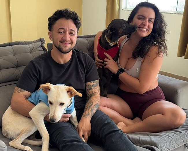 Beni e Camila sorrindo no sofá com as cachorras Sol e Margot